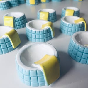 Może przedstawiać: Kolekcja małych, ceramicznych wanien z jasnoniebieskim zewnętrzem i białym wnętrzem. Każda wanna ma żółty ręcznik. Wanny są ułożone na białej powierzchni.