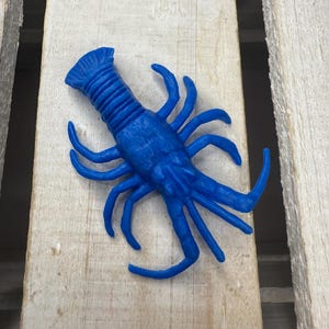 May include: A vibrant blue lobster figurine with detailed claws and segmented body. The crustacean is positioned on a light-colored wooden surface, showcasing its intricate design and bright color.