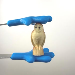 May include: A small, tan owl figurine with brown accents, perched on a wooden base. The owl is held between two blue plastic hand-shaped tools, against a white background. The tools have metal extensions.