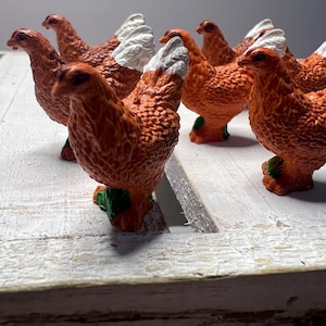 May include: A group of seven orange and white plastic toy chickens. The chickens are standing on a white wooden surface.