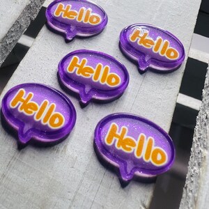 Può includere: Cinque bolle di parole in resina viola con contorno bianco e scritta glitterata arancione che dice "Hello".
