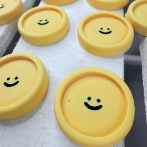 May include: A collection of yellow, circular objects with a smiley face design. The objects are arranged on a white, wooden surface. The objects have a raised edge and a flat top with a black smiley face.