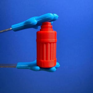 May include: A small, bright orange, cylindrical object with vertical ridges and a stepped top, held between two blue, hand-shaped plastic tongs. The object features a distinct layered texture consistent with 3D printing, set against a solid blue background.