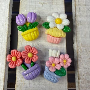 Peut inclure: Quatre décorations de pots de fleurs colorés. Les décorations présentent des fleurs roses, blanches et violettes, avec des centres jaunes et des feuilles vertes. Les pots sont jaunes, roses, violets et bleus. Sur fond de bois.