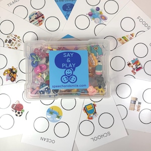 Könnte beinhalten: Ein durchsichtiger Kunststoffbehälter gefüllt mit bunten Miniatur-Anhängern, darunter Schuhe, Bleistifte und Fahrzeuge. Der Behälter hat ein blaues Etikett mit der Aufschrift "SAY & PLAY" und "speechandsmile.com". Weiße Karten mit Kategorien und Kreisen umgeben den Behälter.