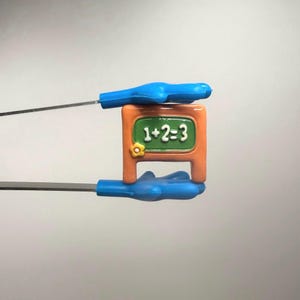 May include: A miniature chalkboard in orange and green, displaying the equation 1+2=3. It's held by blue handles with metal rods. The design is likely for educational purposes.