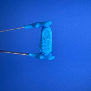 May include: A small, light blue shoe-shaped object is held by a pair of blue tongs against a blue background. The shoe has detailed features, including laces and a sole. The tongs have a silver metal handle.