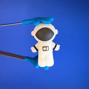 May include: A small, white astronaut figurine with a black visor and black accents. The astronaut is held by blue and silver tools against a solid blue background. The astronaut's suit has a black rectangle with the number 8.