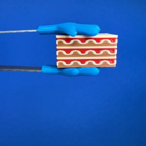 May include: A miniature cake with layers of brown, white, and red frosting is held by two blue-handled tools against a blue background. The cake has a rectangular shape with wavy frosting lines. The tools appear to be cake servers or spatulas.