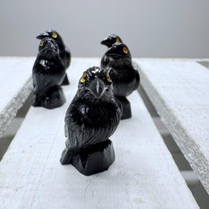 May include: A collection of four glossy black raven figurines with yellow eyes. The birds are perched on small, dark bases, set against a white wooden surface. The ravens are detailed with carved feathers.