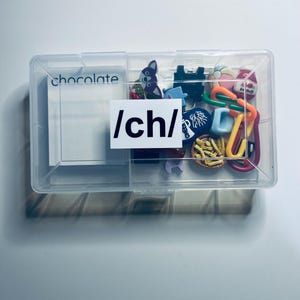 May include: A clear plastic storage box filled with colorful plastic charms and a sticker that says "/ch/". The box also contains a white card with the word "chocolate" written on it.