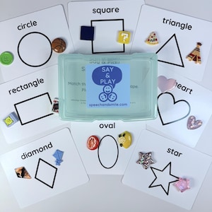 Könnte beinhalten: Lernkarten-Set mit Formen wie Kreis, Quadrat, Dreieck, Rechteck, Raute, Oval, Herz und Stern. Jede Karte zeigt eine Form und ein passendes Objekt. In der Mitte befindet sich eine transparente Kunststoffbox mit dem Text "Say & Play".