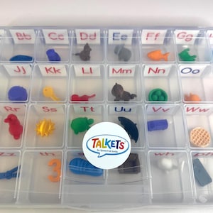 Pode incluir: Um organizador de plástico transparente cheio de pequenos objetos coloridos, cada um em um compartimento separado rotulado com letras do alfabeto. Os objetos incluem formas de animais, alimentos e outros brinquedos pequenos. Um adesivo com a palavra "TALKETS" está no centro.
