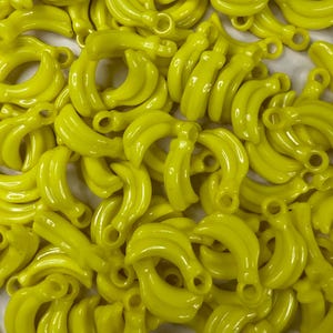 Puede incluir: Un conjunto de dijes amarillos brillantes con forma de plátano. Cada dije tiene un lazo en la parte superior, lo que sugiere que están diseñados para la fabricación de joyas o manualidades. Los dijes son lisos y brillantes, con un color uniforme.