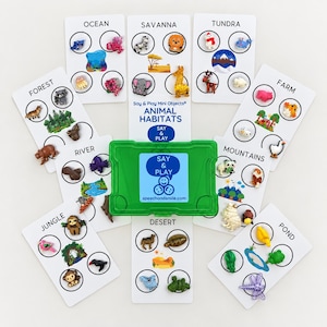 Könnte beinhalten: Ein grüner Kunststoff-Aufbewahrungskoffer mit der Aufschrift Say & Play steht in der Mitte, umgeben von acht weißen Karten, die Tierlebensräume wie Ozean, Savanne, Tundra, Bauernhof, Berge, Teich, Wüste, Dschungel und Wald mit kleinen Tierfiguren zeigen.