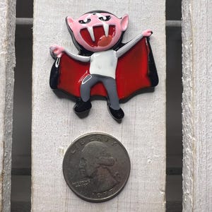 May include: A colorful vampire-themed charm. The charm features a cartoon vampire with a pink face, black hair, and a red cape. The vampire is wearing a white shirt and gray pants. A U.S. quarter is next to the charm.