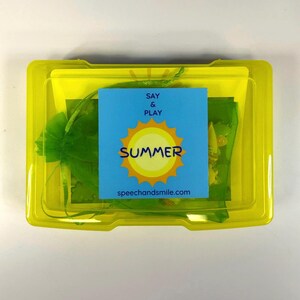 Op de afbeelding: Een transparante gele plastic doos met een blauwe kaart met de woorden "SAY & PLAY SUMMER" en een zonafbeelding. Binnenin zitten groene mesh zakken en andere items. De website speechandsmile.com staat op de kaart.