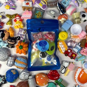 Op de afbeelding: Een blauw hersluitbaar plastic zakje met diverse kleine plastic bedeltjes, voorzien van een ronde sticker met de tekst 'SAY & PLAY'. Het zakje is omringd door een verzameling diverse miniatuurbedeltjes, waaronder een pompoen, een astronaut en een planeet.