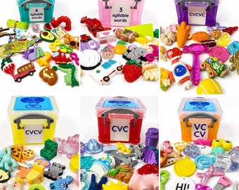 May include: Six colorful plastic storage bins with labels like 4 syllable words, 3 syllable words, CVCVC, CVCV, CVC, and VCCV, each surrounded by a variety of small, multi-colored plastic miniature figurines, charms, and shapes for speech therapy.