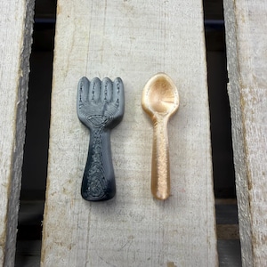 May include: A black and gray fork and a gold spoon are displayed on a wooden surface. The fork has a detailed handle and four tines. The spoon has a rounded bowl and a textured handle. The items appear to be 3D printed.