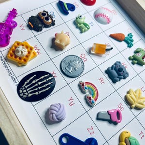 May include: A collection of colorful, miniature charms and embellishments, including a waffle, owl, baseball, and rainbow, arranged on a grid with letters. The charms are made of various materials and feature detailed designs. The wooden frame adds a rustic touch.