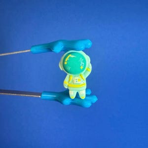 Könnte beinhalten: Eine kleine Astronautenfigur in einem hellgrünen Raumanzug wird von blauen Zangen vor blauem Hintergrund gehalten. Der Astronaut hat einen gelben Helm und winkt. Die Zangen haben blaue Griffe.