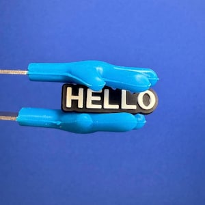 Può includere: Primo piano di un ciondolo per scarpe blu con la scritta "HELLO" in lettere bianche su sfondo nero. Il ciondolo è circondato da due accessori blu a forma di dito, il tutto su uno sfondo blu.