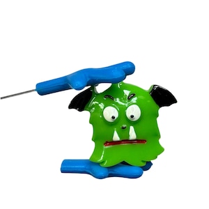 May include: A green monster-shaped pin with white eyes, a red mouth, and black ears. The pin is held by a blue plastic handle and a thin metal needle. The design is cartoonish and playful.