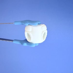 May include: A white, rounded object with four openings is held by two blue-handled tools against a blue background. The object has a textured surface and a cylindrical shape. The tools appear to be surgical instruments.