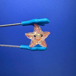 Puede incluir: Un dije de estrella de mar sonriente y translúcida con un interior brillante es sostenido por unas pinzas azules sobre un fondo azul. La estrella de mar tiene una cara feliz con ojos negros y una sonrisa roja. El dije mide aproximadamente 3,8 cm de ancho.