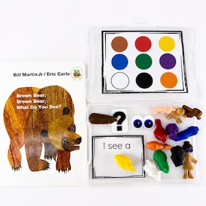 May include: A Brown Bear, Brown Bear, What Do You See? book by Bill Martin Jr and Eric Carle next to a clear plastic storage case containing a color-coded grid, a question mark tile, two googly eyes, a card reading I see a, and small figurines.