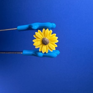 Peut inclure: Une fleur de tournesol jaune vif avec un centre marron est tenue par deux outils à poignée bleue sur un fond bleu. Le tournesol est au centre de l'attention, avec ses pétales et son centre détaillés. Les outils ressemblent à des pinces.