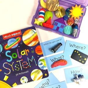 May include: A Hello, World! Solar System book by Jill McDonald, a purple plastic storage case filled with colorful small figurines including a sun, house, and rocket, and several light blue cards with questions and small objects attached.