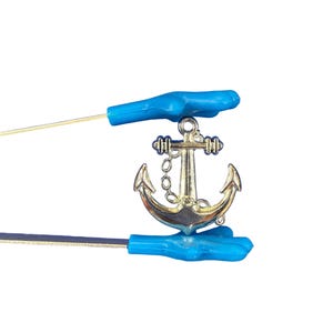 Puede incluir: Un dije de ancla de color dorado con una cadena, sostenido por dos herramientas azules. El ancla tiene un diseño clásico, con un poste central y brazos curvos. Las herramientas tienen mangos azules y vástagos finos de color plateado.