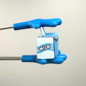 Puede incluir: Un pequeño cartón de leche azul claro con la palabra "MILK" y estrellas. Lo sostienen manos de plástico azul. El cartón es blanco con un diseño azul.