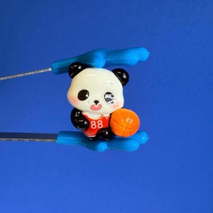 May include: A panda figurine wearing a red basketball jersey with the number 88, holding an orange basketball. The panda has black ears, eyes, and arms, with a white face. The background is a solid blue.