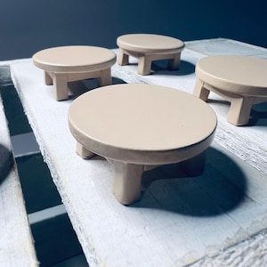 May include: Four small, round, beige wooden tables with four legs each. The tables are arranged on a white wooden surface.