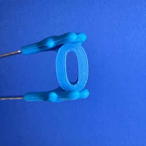 May include: A blue number zero is held by a pair of blue-handled tweezers against a blue background. The number is made of a solid material and has a textured surface. The tweezers are silver in color.
