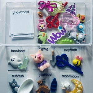 Op de afbeelding: Een set van 10 kleine plastic kaartjes met woordparen erop gedrukt. Elk kaartje heeft een ander kleurrijk plastic sieraad eraan bevestigd. De sieraden omvatten een ​​spook, een laars, een varken, een schop, een sleutel, een bij, een beer, een badkuip, een geit, een boot, een koe en een wassende maan.