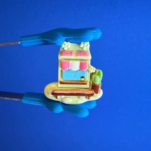 Peut inclure: Une figurine miniature de magasin colorée avec un extérieur vert clair, des accents roses et un auvent bleu. La figurine comprend un petit banc, un cactus en pot et une petite tasse de café jaune. Le fond est bleu uni.