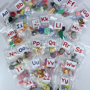 May include: A selection of small, colourful embellishments, each in a clear plastic bag labelled with a letter of the alphabet. The bags contain various shapes and designs, including fruits, animals, and other decorative items.