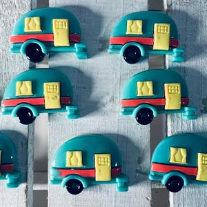 May include: A collection of teal camper trailer-shaped embellishments. Each trailer has a red stripe, yellow door and windows, and black wheels. The trailers are arranged on a white, textured surface.