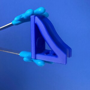 May include: A small, dark blue, triangular-shaped plastic component with a curved slide-like feature on one side and a flat vertical edge. The object is held by a pair of metal tweezers with bright blue, hand-shaped silicone tips.