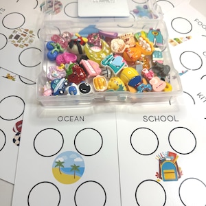 Könnte beinhalten: Eine transparente Kunststoffbox gefüllt mit bunten Miniatur-Anhängern, darunter Lebensmittel, Tiere und Objekte. Die Box liegt auf weißen Karten mit den Aufschriften "Ocean" und "School" mit Kreisen für die Anhänger.