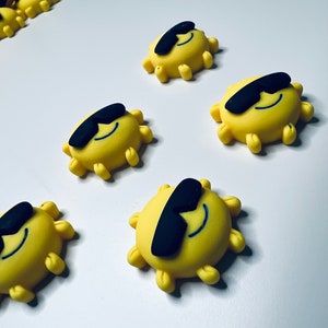 May include: Six yellow sun-shaped decorations with black sunglasses. The suns have a smiling face and are made of a soft, pliable material.