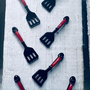 May include: Miniature black spatulas with red handles are arranged on a white, weathered wooden surface. The spatulas have a slotted design on the spatula head. The red handles have a rounded black top.