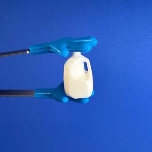 May include: A small, off-white plastic milk jug with a handle is held by blue-gloved hands against a blue background. The jug has a rectangular shape with a curved top and a small opening.
