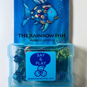 May include: A blue plastic container with a clear lid filled with colourful plastic toys. The container has a blue sticker that says "Say & Play" with a speech bubble and three smiley faces. The text "speechandsmile.com" is below the smiley faces. The container is sitting on top of a blue book with a picture of a rainbow fish and the title "The Rainbow Fish" by Marcus Pfister.