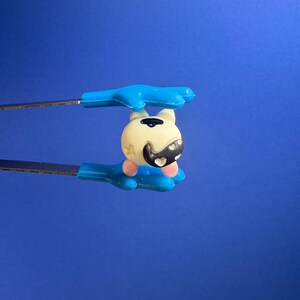 May include: A small, round, cream-colored bead featuring a black cat-like face design with pink accents, held securely between the blue, hand-shaped tips of a pair of metal tweezers against a solid, vibrant blue background.