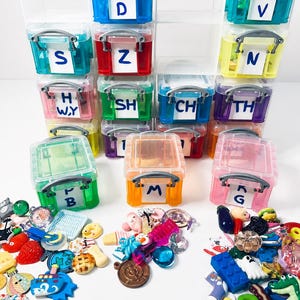 May include: A collection of small, colorful, translucent plastic storage containers with silver handles, each labeled with white paper tags featuring black letters or letter combinations. The containers are filled with various miniature charms.